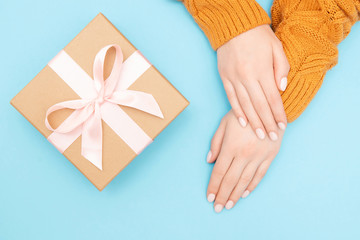 Obraz na płótnie Canvas Beautiful female hands with pink manicure on nails in warm knitted sweater blue background with gift craft box baht. Concept present Valentines day, Birthday, Christmas