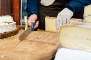 Close up on midsection hands of the man sale selling the french cheese saint nectaire holding big knife on the board at his store cutting cut in the pieces