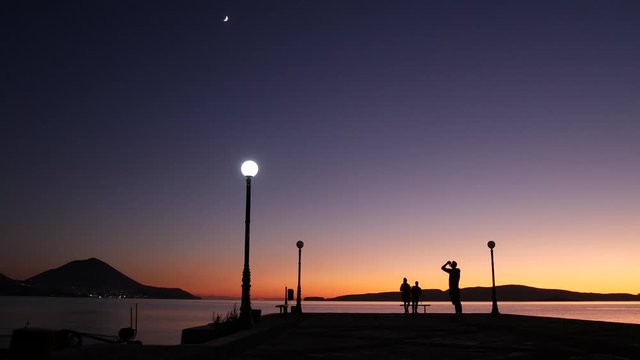 Epic shot of a purple sunset view from a pier. People watching and taking pictures. The moon is appearing on the night sky. Beautiful and romantic atmosphere.
