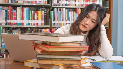 Close up young asian woman siting and thinking seriously while looking stack of book on desk in library with bookshelf background prepare exam university or school, education and searching  concept.