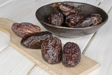 Group of eight whole dried brown date fruit on wooden cutting board in glazed bowl on white wood