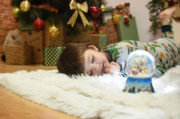 Fototapeta na wymiar Boy looking a glass snow ball under a Christmas tree. Child with a snow ball with a New Year