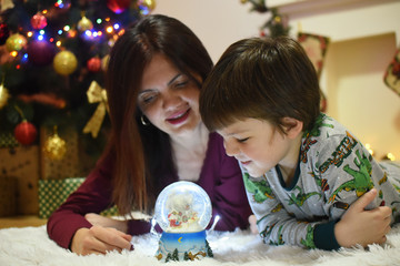 Mom and little boy looking a glass snow ball under a Christmas tree. Mother and son with a snow ball with a New Year