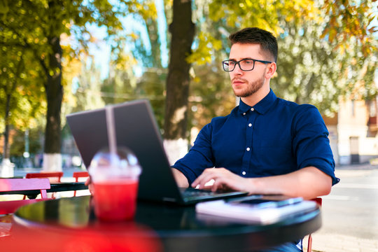 Young Attractive Hipster Freelancer Working Remotely On Laptop At Outdoor Cafe. Businessman Browsing Internet At Coffeshop. Blogger Posts To Social Media On Travel Using 5g Internet. Telecommuting.