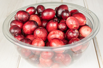 Lot of whole fresh red cranberry in round transparent plastic bowl on white wood