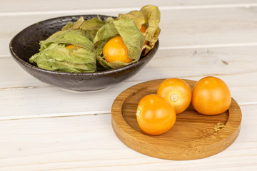 Lot of whole fresh orange physalis in glazed bowl on round bamboo coaster on white wood