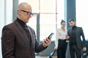 Naklejka na ściany i meble Serious bald mature businessman in formalwear looking through data in smartphone
