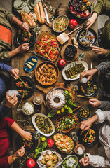 Traditional Turkish celebration dinner at rustic table. Flat-lay of people eating Turkish salads, cooked vegetables, meze starters, pastries and drinking raki drink, top view. Middle Eastern cuisine