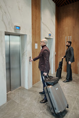One of businessmen with suitcases pushing call button of elevator in hotel