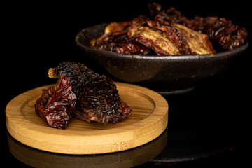 Lot of whole dried red tomato piece in glazed bowl on round bamboo coaster isolated on black glass