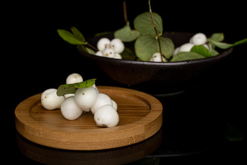 Lot of whole white snowberry on round bamboo coaster in glazed bowl isolated on black glass