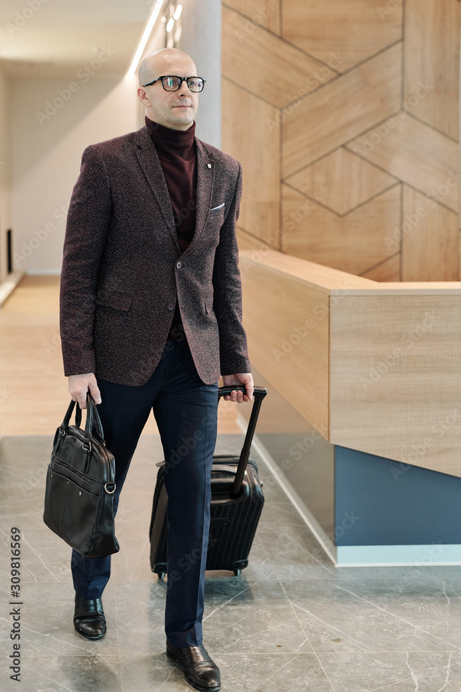 Poster bald mature businessman pulling suitcase while moving along reception counter