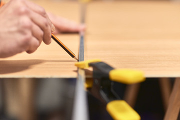 Detail of a man's hand making a mark with a pencil and a metal guide on a wooden board. The guide is held in place with a clamp.