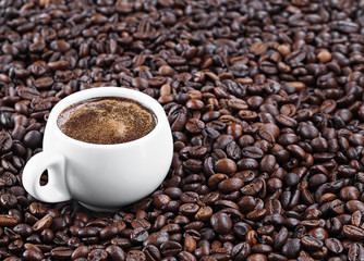 A white cup with aromatic espresso stands in the coffee beans. Roasted coffee beans, background. Horizontal orientation. Fresh aromatic dark coffee. Close-up. Selective focus on a cup with espresso.