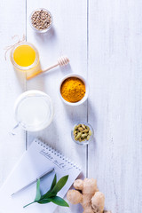 Ingredients for Golden Latte on a white wooden background. Ginger root, milk, turmeric, honey, cardamom, allspice peas and a notebook with a clean sheet.
