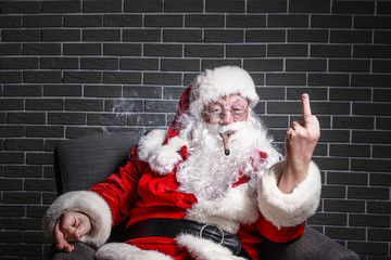 Funny drunk Santa Claus with cigar showing middle finger while sitting in armchair against brick...