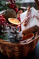 Wicker basket with striped candy canes, dried sliced oranges, cones and gifts. Cozy atmosphere at home before Christmas. Rustic basket with green spruce branches and gifts in craft paper.