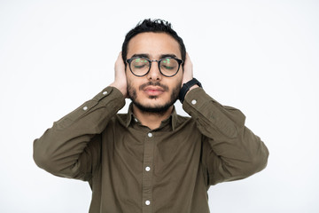 Young annoyed man in eyeglasses keeping his eyes closed and hands on ears