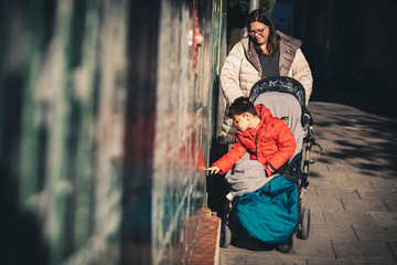 Fototapeta na wymiar una mujer (la madre) lleva a su hijo con pluridiscapacidad de paseo por la calle en invierno con cochecito adaptado (silla de ruedas), el niño se acerca al muro para tocarlo con la mano, estimulo sens