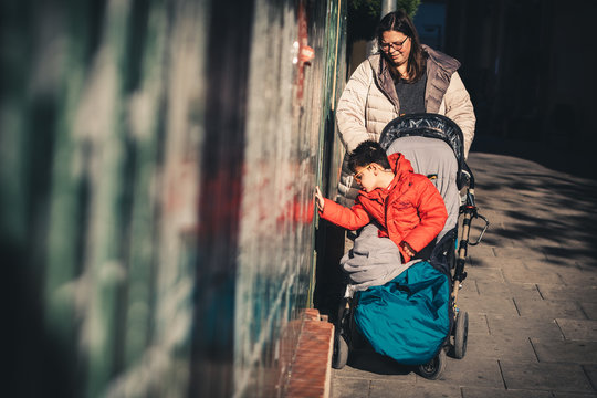 una mujer (la madre) lleva a su hijo con pluridiscapacidad de paseo por la calle en invierno con cochecito adaptado (silla de ruedas), el ni&ntilde;o se acerca al muro para tocarlo con la mano, estimulo sens