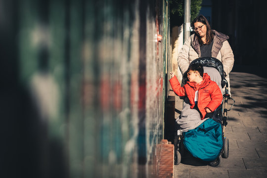 una mujer (la madre) lleva a su hijo con pluridiscapacidad de paseo por la calle en invierno con cochecito adaptado (silla de ruedas), el ni&ntilde;o se acerca al muro para tocarlo con la mano, estimulo sens