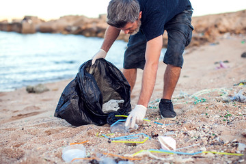 Man with white gloves and big black package collecting garbage on the beach. Environmental protection and planet pollution concept