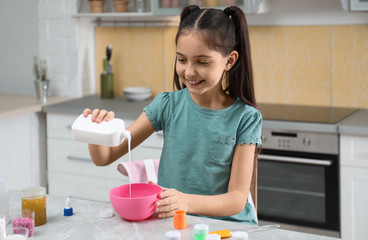 Cute little girl pouring glue into bowl at table in kitchen. DIY slime toy