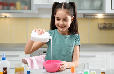 Cute little girl pouring glue into bowl at table in kitchen. DIY slime toy