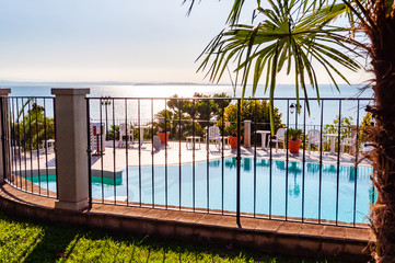 Outdoor pool behind the fence with crystal water, parasols and deck chairs with Garda lake and Sirmione city on the background