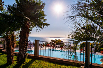 Sunny outdoor pool behind the fence with crystal water, parasols and deck chairs with Garda lake and Sirmione city on the background