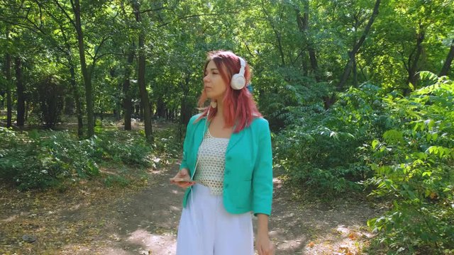 Young happy woman in colorful suit and with pink hair walk in summer park and listen to music