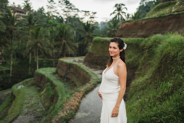 Young pregnant woman in white dress with view of Bali rice terraces in morning sunlight. Harmony with nature. Pregnancy concept.