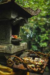 Traditional balinese Canang Sari offerings to gods and spirits with flowers, food and smoky aromatic sticks on dark green background. Indonesian culture and religion. Bali authentic travel concept.