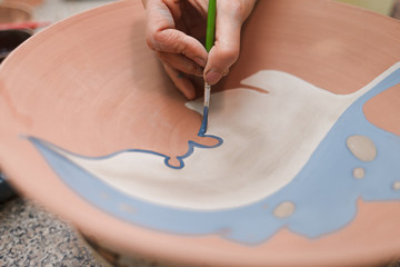 Potter woman paints a ceramic plate closeup. Girl draws with a brush on earthenware. Process of creating clay products. 