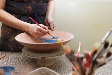 Fototapeta na wymiar Potter woman paints a ceramic plate. Girl draws with a brush on earthenware. Process of creating clay products