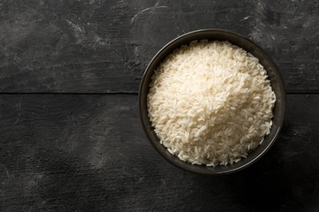 White uncooked, raw long grain rice in black bowl on rustic, dark wooden table with copy space top view flat lay from above