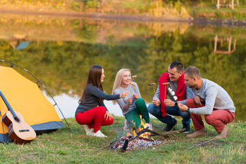 travel, tourism, hike, picnic and people concept - group of happy friends frying sausages on campfire near lake.