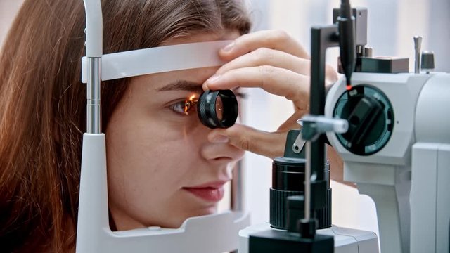 Ophthalmology treatment - a young woman with pink lips checking her visual acuity with a light of special big optometry machine and lens - brown eyes color