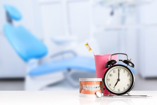 Dental mirror and dental explorer instrument near human jaw, alarm clock and toothbrush in pink glass against dental office chair background. Brush teeth concept.