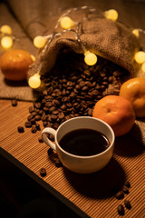 Holiday still life with a cup of brewed coffee, coffee grains and tangerines. The lights of the garland give comfort. Vertical photo