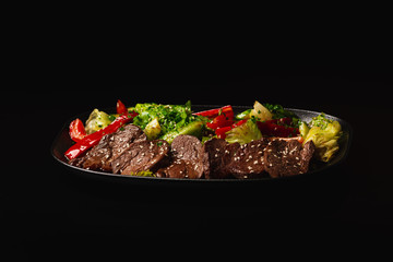 Juicy Beef Sirloin Steak Salad with bell pepper, sesam seeds and green vegetables in a black plate, on black background. Healthy food