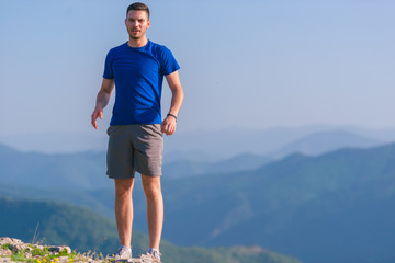 Tall male wearing sports clothes standing on top of a cliff while pointing at something.