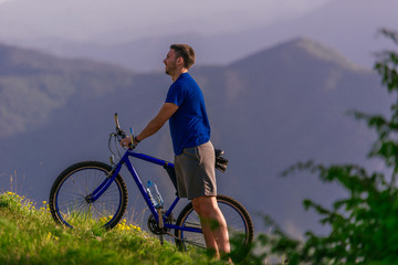 Tired cyclist is wiping his sweat off his face while pushing his bicycle uphill on a dirt road in a mountain.