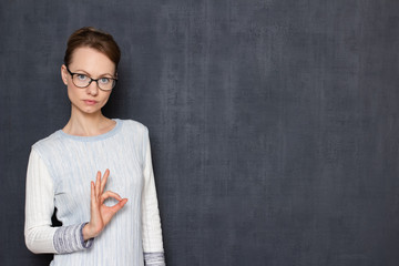 Portrait of serious young woman with glasses, showing ok gesture