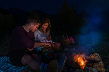 Young couple baking sausages on the campfire and drinking beer in the forest hill in the dusk