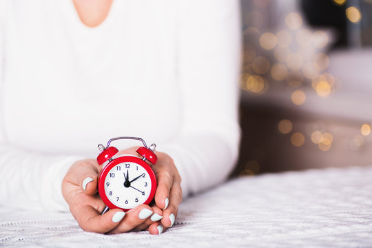 Female Hands Holding Little Red Alarm Clock With Warm Garland Bokeh On Blurred Background Indoor.