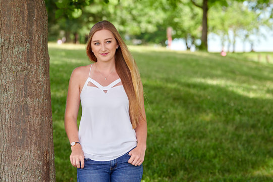 Stunning young blonde caucasian woman in white blouse and blue jeans poses in park near tree