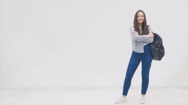 Happy, smiling, full height confident girl with long hair appearing on a white background with backpack slung over her shoulder, and is folding her hands.