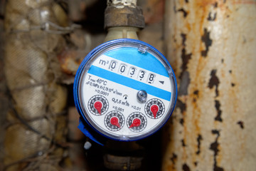 Blue cold water meter against the background of a rusty pipe.  Indoor measurement of water flow.