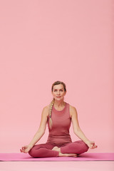 Fototapeta premium Full length portrait of fit mature woman doing yoga against pastel pink background, lotus position